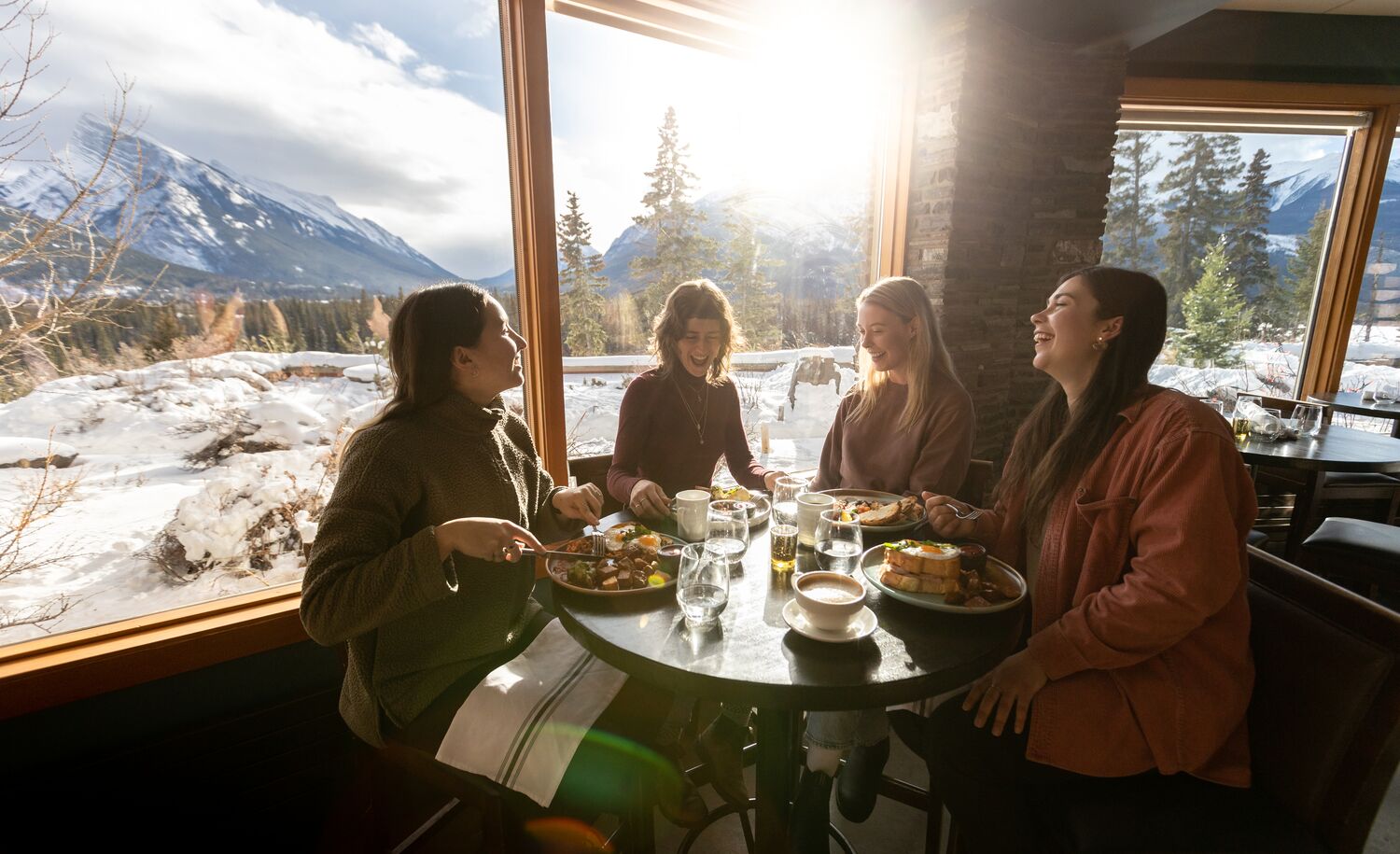Four friends sit at a table eating brunch with mountains out the window behind them at the Juniper Bistro in Banff.